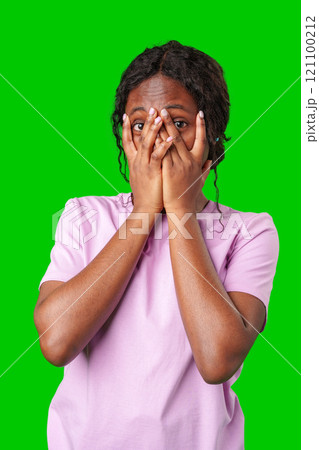 A young woman in a pink shirt covers her face with both hands against green background 121100212