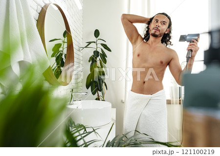 A man with curly hair styles himself in a bright room while using a hairdryer to enhance his look A man with curly hair styles himself in a bright room while using a hairdryer to enhance his look 121100219