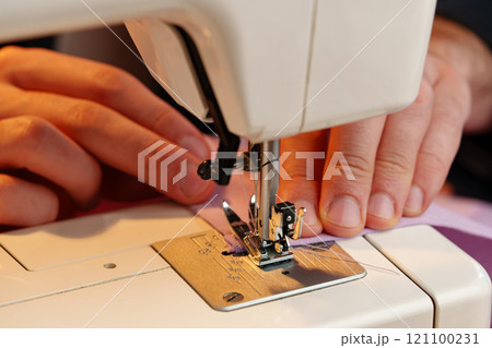 Hands sewing fabric using a sewing machine on a wooden table in a well-lit workspace Hands sewing fabric using a sewing machine on a wooden table in a well-lit workspace 121100231