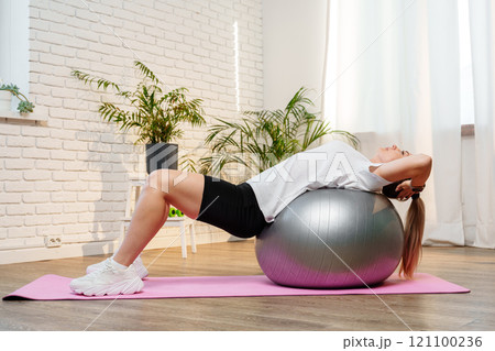 Young woman exercising on a stability ball in a bright room with plants Young woman exercising on a stability ball in a bright room with plants 121100236