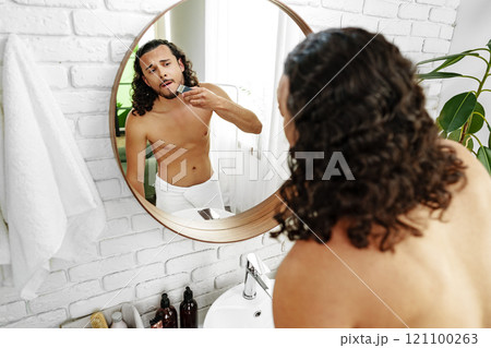 A man with curly hair grooming himself in the bathroom while looking into a round mirror 121100263