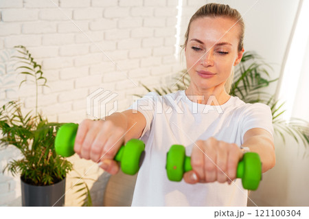 A woman exercises with green dumbbells at home in a bright room 121100304