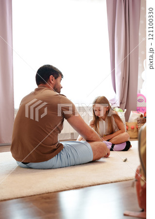 A father and his daughter sit on a rug in their home, the father looking at his daughter with a gentle expression A father and his daughter sit on a rug in their home, the father looking at his daughter with a gentle expression 121100330