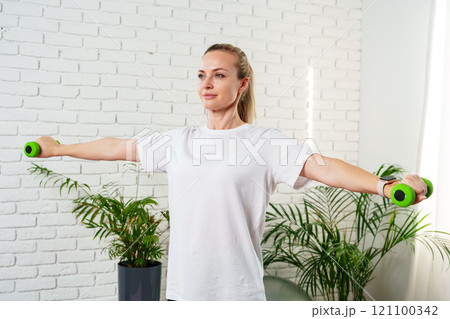 A woman exercises with green dumbbells at home in a bright room A woman exercises with green dumbbells at home in a bright room 121100342