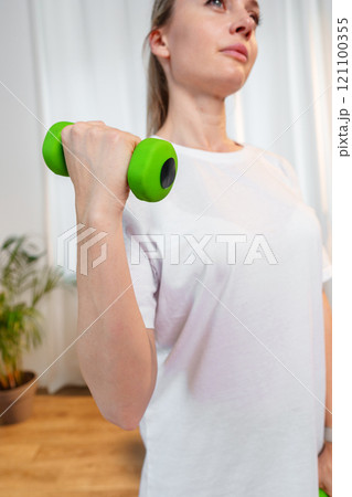 A woman exercises with green dumbbells at home in a bright room 121100355