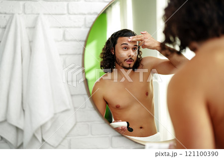 A young man applies skincare cream while looking in a round mirror in a bright bathroom A young man applies skincare cream while looking in a round mirror in a bright bathroom 121100390