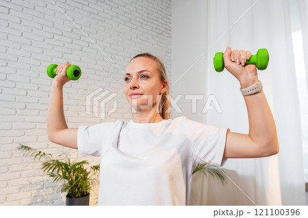 A woman exercises with green dumbbells at home in a bright room 121100396