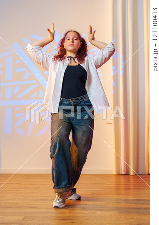 A young woman dances in a studio, her arms raised above her head, with a white shirt and blue jeans 121100413