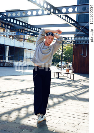 A young man in a grey sweatshirt and black pants stands in an urban setting, his arms crossed behind his head 121100554