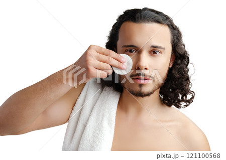 A young man with curly hair applies a skincare product to his face in a well-lit space 121100568