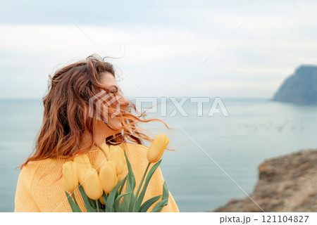 Portrait of a happy woman with hair flying in the wind against the backdrop of mountains and sea. Holding a bouquet of yellow tulips in her hands, wearing a yellow sweater Portrait of a happy woman with hair flying in the wind against the backdrop of mountains and sea. Holding a bouquet of yellow tulips in her hands, wearing a yellow sweater 121104827