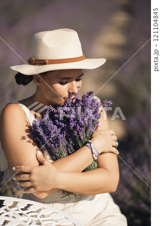 woman sitting in a field of lavender and wearing a straw hat. She is smiling and holding a bouquet of flowers. Scene is peaceful and serene, as the woman is surrounded by the beauty of nature 121104845