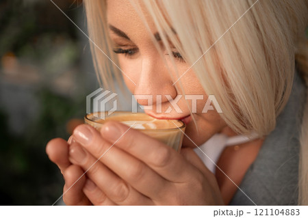 A blonde woman is drinking coffee from a glass. She is smiling and she is enjoying her drink. 121104883