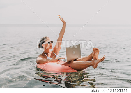 A woman is sitting on a pink inflatable raft in the ocean, holding a laptop. She is smiling and she is enjoying her time. Concept of relaxation and leisure. A woman is sitting on a pink inflatable raft in the ocean, holding a laptop. She is smiling and she is enjoying her time. Concept of relaxation and leisure. 121105531
