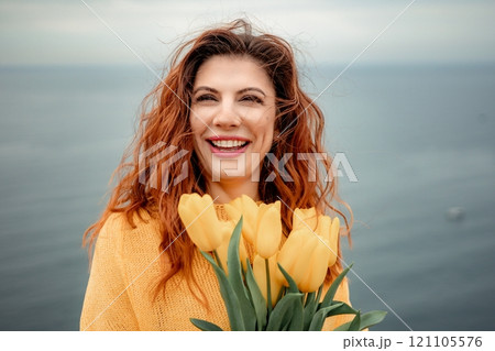 Portrait of a happy woman with hair flying in the wind against the backdrop of mountains and sea. Holding a bouquet of yellow tulips in her hands, wearing a yellow sweater 121105576