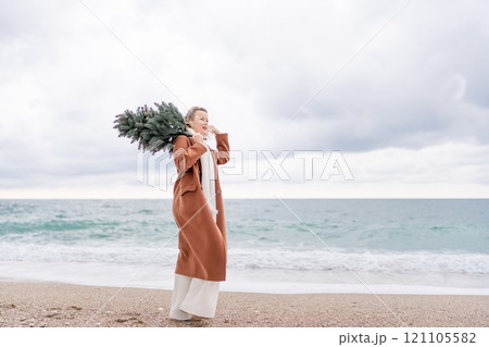 Blond woman Christmas tree sea. Christmas portrait of a happy woman walking along the beach and holding a Christmas tree on her shoulder. She is wearing a brown coat and a white suit. 121105582