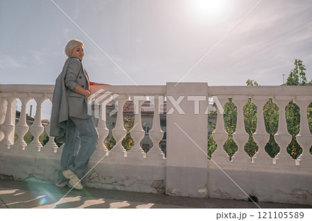 A woman in a gray jacket and blue jeans is leaning against a white railing. She is holding a book in her hand. The scene has a calm and relaxed mood, with the woman enjoying a quiet moment outdoors. 121105589
