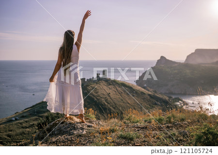 A woman in a white dress stands on a rocky hill overlooking the ocean. She is smiling and she is happy. 121105724
