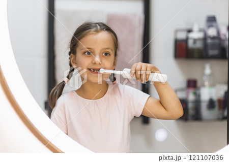 Little Caucasian girl with lost baby teeth using electric toothbrush, looking in bathroom mirror, promoting dental hygiene. 121107036