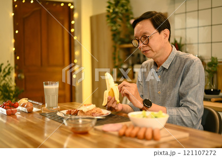 Happy senior man man sitting at wooden kitchen counter, peeling a banana 121107272
