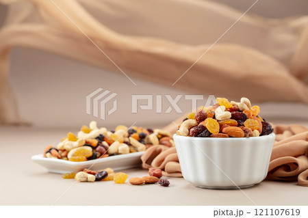 The mix of various nuts and raisins in a white bowl on a beige background. The mix of various nuts and raisins in a white bowl on a beige background. 121107619