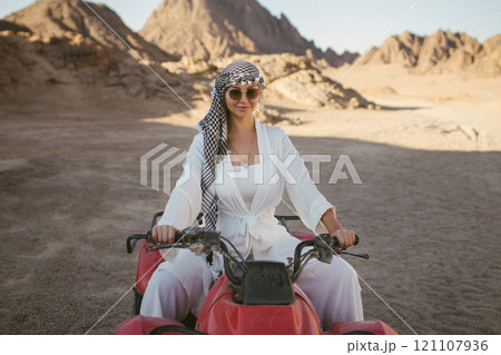 A rather attractive woman is going on a safari, driving an ATV in the desert. Beautiful young woman with a scarf on her head sits on a red four-wheeled ATV and rides towards adventure 121107936