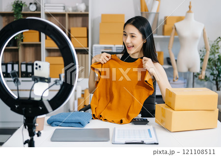 Fashion blogger concept, Young Asian women selling clothes on video streaming.Startup small business SME, using smartphone or tablet taking receive and checking 121108511