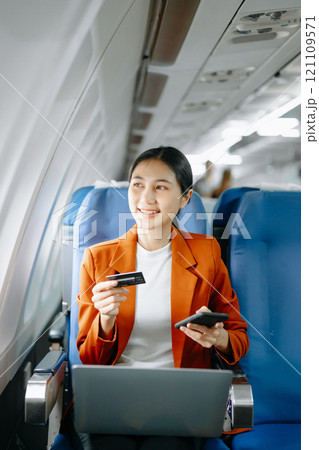 Asian businesswoman or entrepreneur in a formal suit on an airplane sitting in business class using a phone, computer laptop. Travel in style, work 121109571
