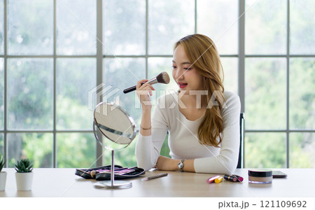 A woman is happily applying makeup while looking in a mirror at a table with various beauty products. 121109692
