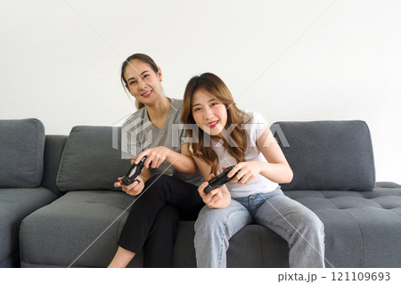 Two asian women playing console game, sitting on sofa in the living room. Enjoy leisure weekend at home. Stress free concept. 121109693