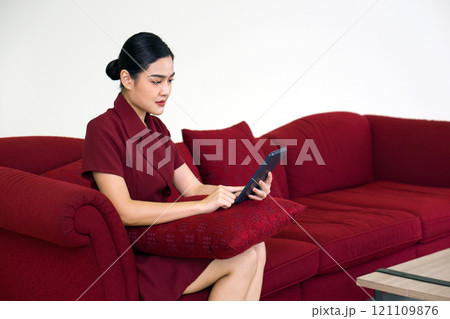 Young asian businesswoman in red dress working on red sofa at home. Tablet computer and cushion are on her lap. Young asian businesswoman in red dress working on red sofa at home. Tablet computer and cushion are on her lap. 121109876