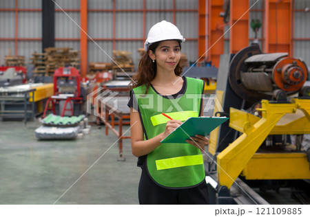 A woman wearing a safety vest and hardhat stand in a factory. She holds a clipboard and looks confident. A woman wearing a safety vest and hardhat stand in a factory. She holds a clipboard and looks confident. 121109885