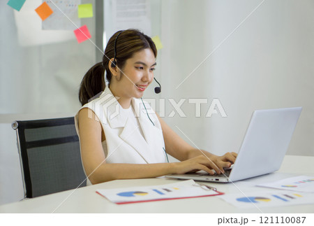 Young asian businesswoman in  headset with mic typing on laptop computer keyboard while meeting online through internet connection. Working concept social distancing. 121110087