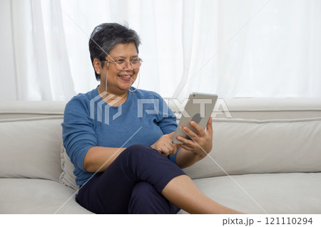 Asian gray hair old woman looking at tablet computer monitor, smiling while sit on sofa in the living room. 121110294