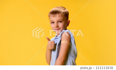 Little boy posing demonstrating patch on hand and gesturing thumb up after vaccination against yellow background. 121111616