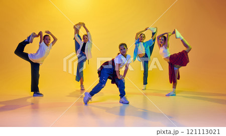 Flexibility and talent. Group of teen girls in casual stylish attire in motion, dancing hip hop together against yellow background in neon light Flexibility and talent. Group of teen girls in casual stylish attire in motion, dancing hip hop together against yellow background in neon light 121113021