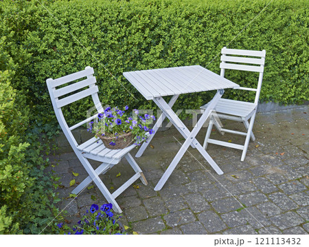 Two courtyard chairs and table in a secluded and private garden at home. Wooden patio furniture and seating area for coffee and reading in a backyard. Purple meadow cranesbill flowers and a hedge 121113432
