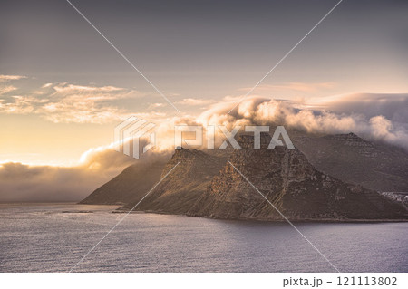Panorama of a mountain coastline with a cloudy sunset in South Africa. Scenic landscape of soft white clouds covering Table Mountain at dusk near a calm peaceful sea at Hout Bay near Cape Town. 121113802
