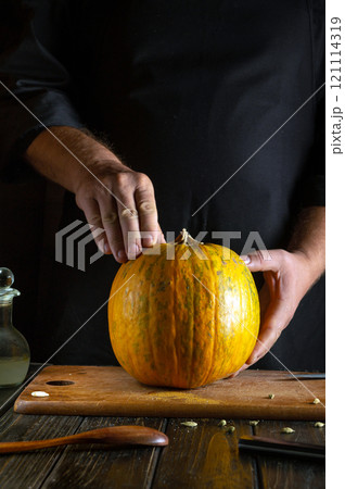 A skilled chef gently inspects a bright orange pumpkin resting on a wooden cutting board, surrounded by kitchen tools and an olive oil bottle, preparing for a seasonal recipe A skilled chef gently inspects a bright orange pumpkin resting on a wooden cutting board, surrounded by kitchen tools and an olive oil bottle, preparing for a seasonal recipe 121114319