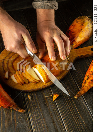 Hands skillfully slice vibrant sweet beetroot on a beautifully grained wooden board. The warm tones of the kitchen create an inviting atmosphere for cooking 121114325