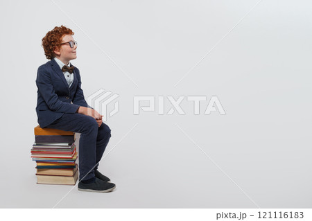 Smiling redhead child schoolboy sitting on stack of books and looking up against white studio wall Smiling redhead child schoolboy sitting on stack of books and looking up against white studio wall 121116183