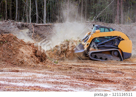 Heavy bulldozer machinery operates to clear section of land, creating dust clouds as it churns through dirt in construction site near at forested area 121116511