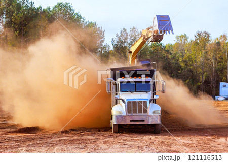 Construction truck is unloading dirt while large excavator scoops material dust clouds billow indicating active earthmoving operations 121116513