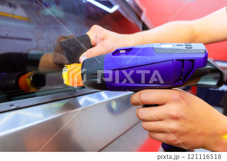 Technician is using heat gun to carefully remove window tinting from vehicle side window in car workshop. 121116518