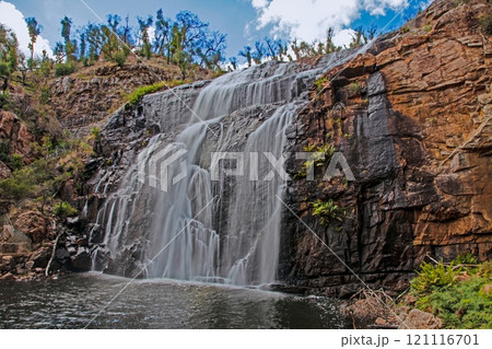 Waterfall cascading down rugged cliffs surrounded by lush greenery under a bright blue sky 121116701