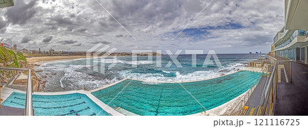 Panoramic view of Bondi Beach in Sydney, Australia, featuring the iconic Bondi Icebergs pool and ocean waves under a cloudy sky 121116725