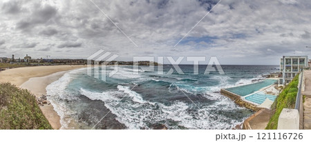 Panoramic view of Bondi Beach in Sydney, Australia, featuring the iconic Bondi Icebergs pool and ocean waves under a cloudy sky 121116726