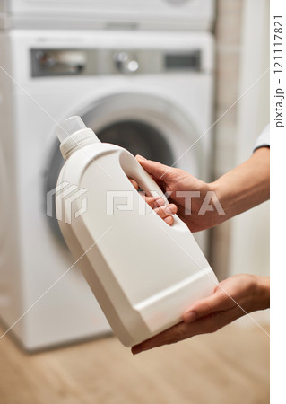 female hands hold white bottle with laundry gel or liquid softener against blurred washing machine at home. female hands hold white bottle with laundry gel or liquid softener against blurred washing machine at home. 121117821