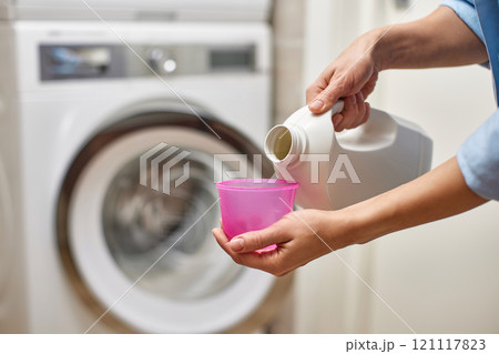female hand pours liquid transparent laundry detergent or conditioner into pink cap against washing machine. female hand pours liquid transparent laundry detergent or conditioner into pink cap against washing machine. 121117823