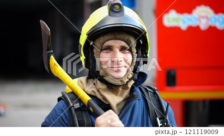 Portrait of happy male fireguard with axe standing near a fire engine. Young smiling fireman looking at camera in full equipment after fire near big red truck. Saving lives and heroic profession Portrait of happy male fireguard with axe standing near a fire engine. Young smiling fireman looking at camera in full equipment after fire near big red truck. Saving lives and heroic profession 121119009